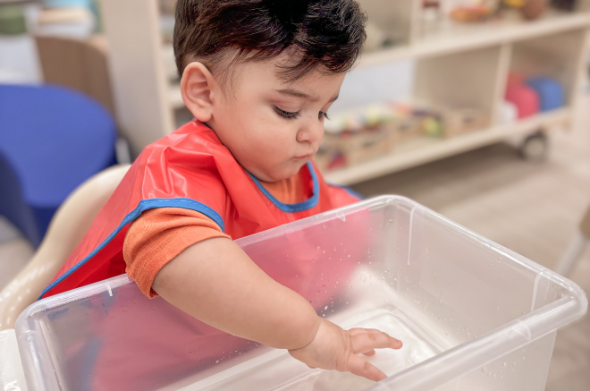 Young child in a red apron reaches into a clear plastic tub of water at a play area indoors.
