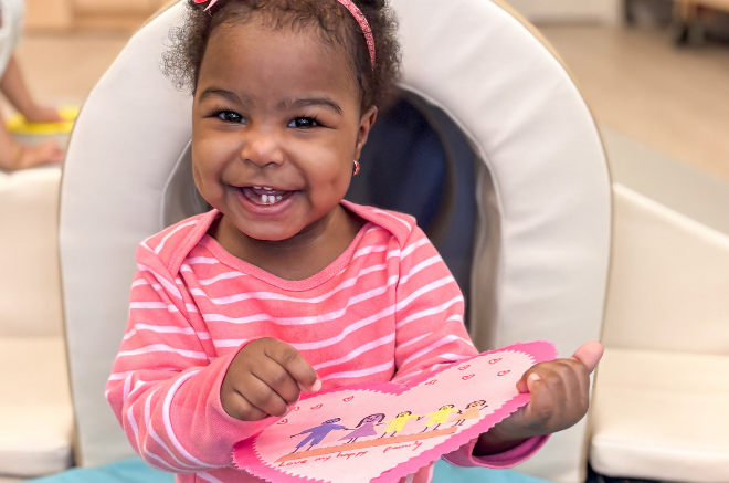 Smiling toddler girl in a pink striped shirt sits in a cushioned chair, proudly holding a pink paper craft with drawings.
