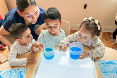 Teacher guides three preschoolers as they explore water at a light table with blue cups on the table edge behind them.