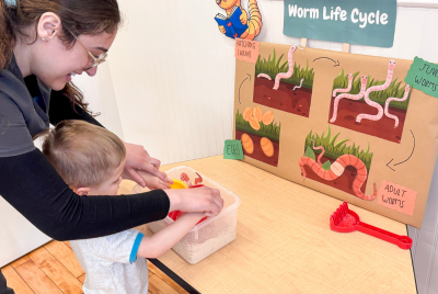 Caregiver guiding a toddler's hands as they explore a worm life cycle display on a classroom table.