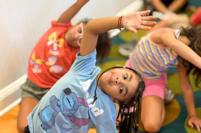 Children in a classroom doing a side stretch; foreground girl with braided hair in a blue shirt reaches up and to the side, others stretching nearby.