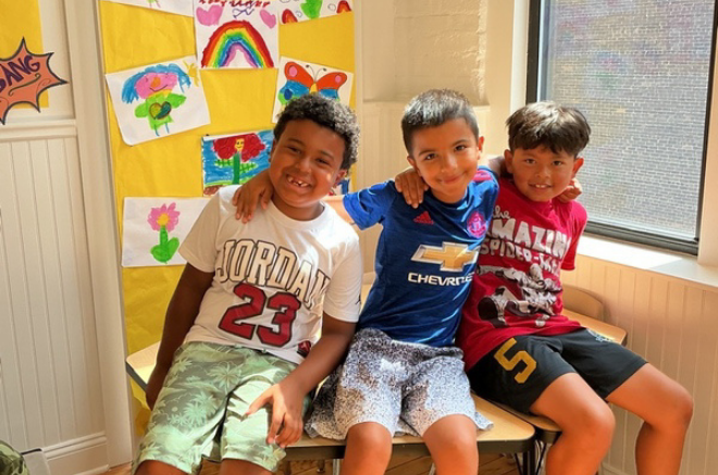 Three smiling boys sit side by side with arms around each other in a bright room, colorful kids’ drawings on a yellow display behind them.
