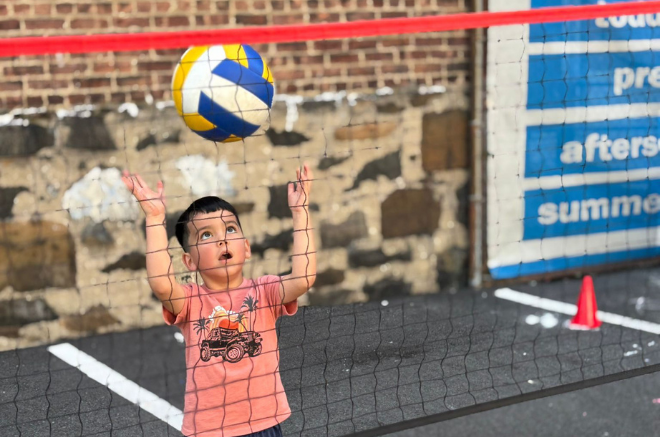 Young child in a pink shirt reaches up to hit a volleyball over a net on an outdoor court, with a brick wall in the background.