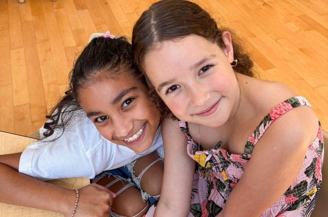 Two young girls smiling and leaning their heads together on a wooden floor; one wears a white shirt, the other a floral dress.
