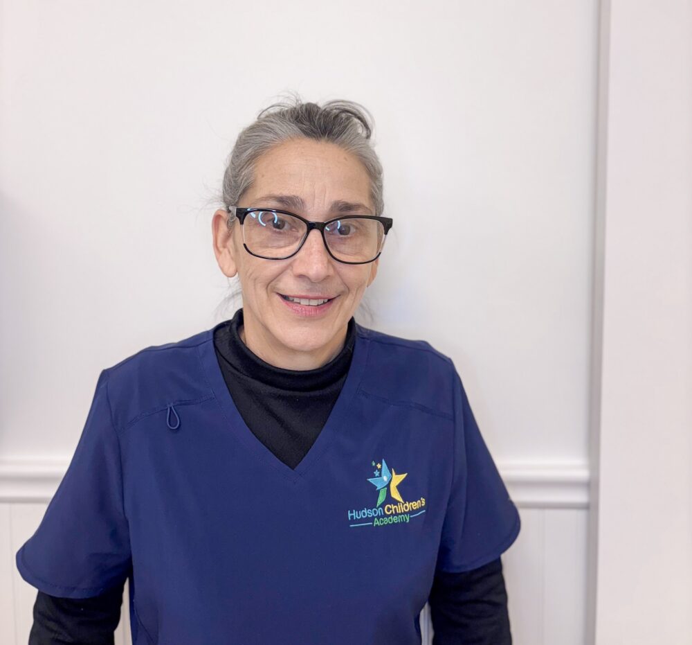 Smiling woman with gray hair and glasses in blue scrubs, standing against a light wall; logo says Hudson Children's Academy on her chest.