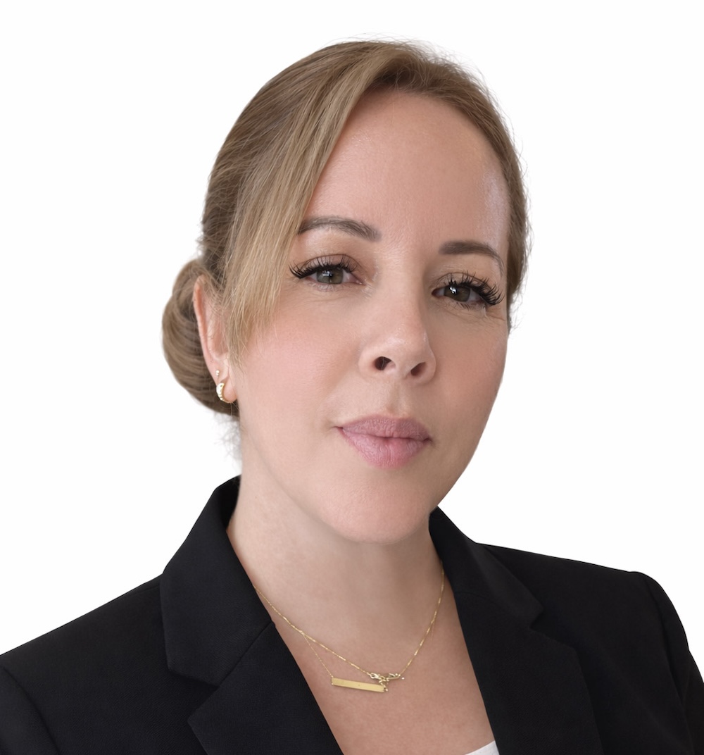 Portrait of a woman with blonde hair tied in a bun, wearing a black blazer and a gold necklace against a white background.