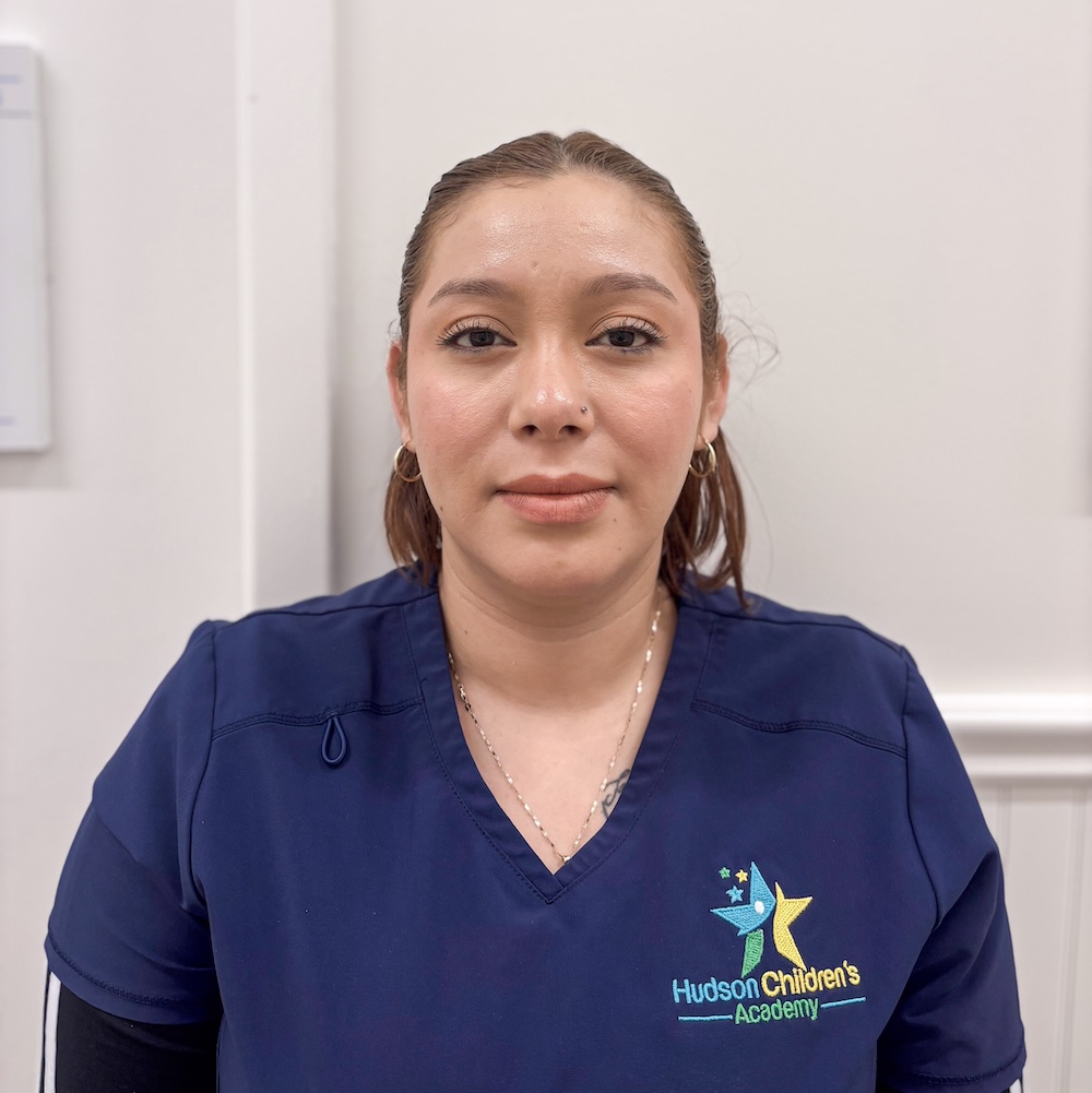 Head-and-shoulders portrait of a woman in navy scrubs with the Hudson Children's Academy logo, looking at the camera in a clinical setting.