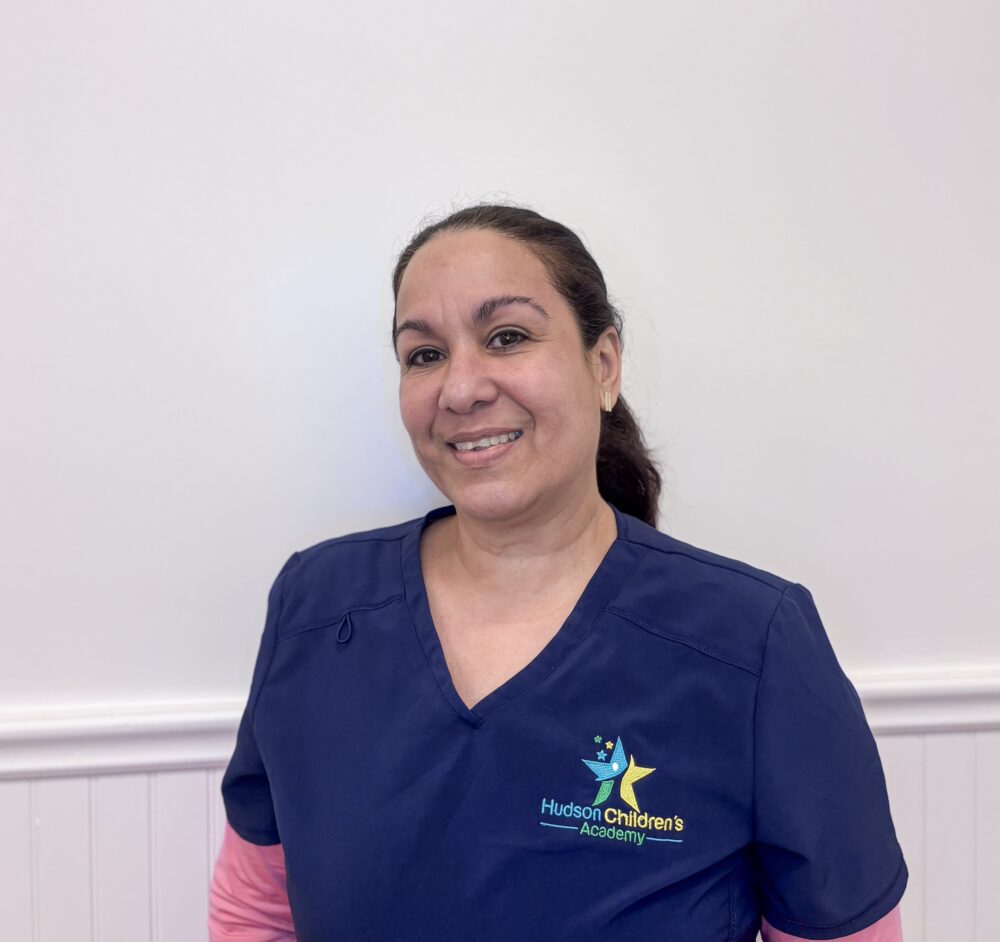 Smiling woman in navy scrubs standing against a pale wall, with Hudson Children's Academy logo on her chest.