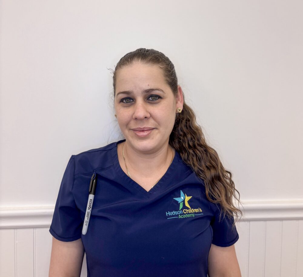 Portrait of a woman in navy scrubs with a Hudson Children’s Academy logo, standing against a pale wall. A lanyard and marker are visible on her chest.