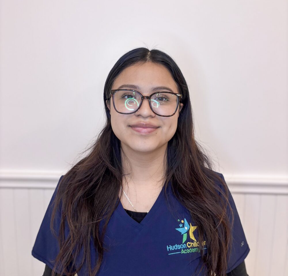 Young woman in navy scrubs with embroidered star logo and 'Hudson Child Academy' text, standing against a light wall.