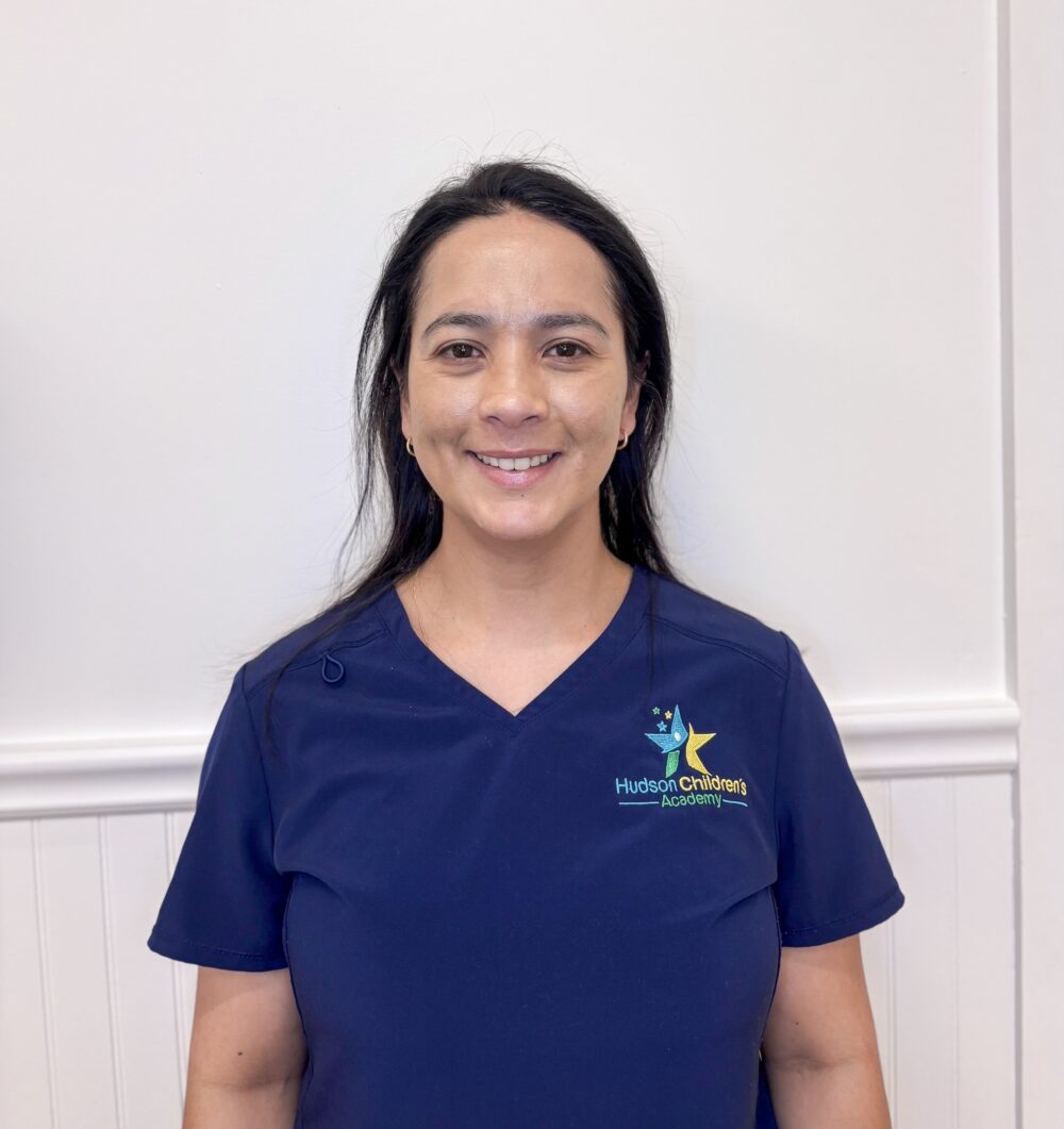 Smiling woman in navy scrubs with Hudson Children's Academy logo against a white wall.