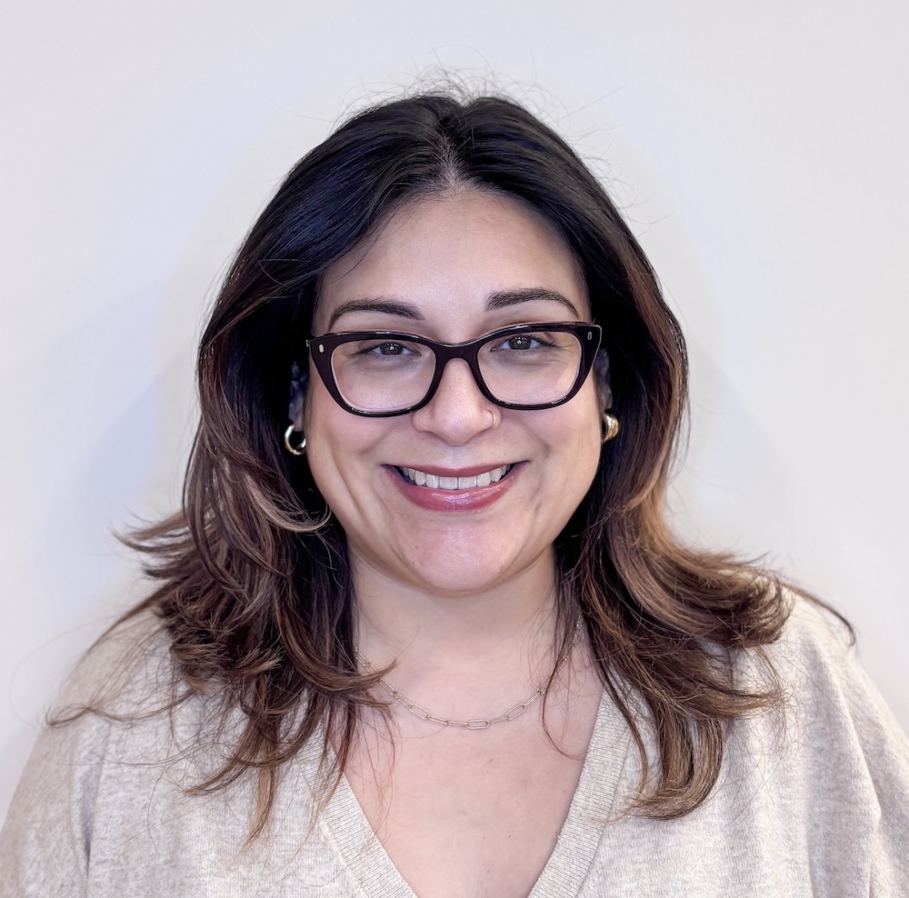 Smiling woman with dark hair and glasses, wearing a light beige sweater and gold hoop earrings, facing the camera.
