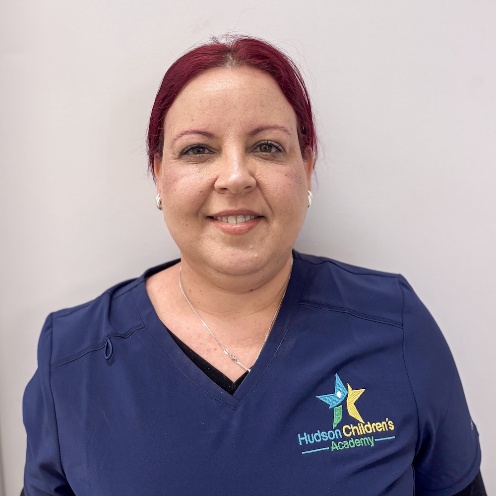 Smiling woman in navy scrubs with Hudson Children's Academy logo on the chest, standing against a gray background.