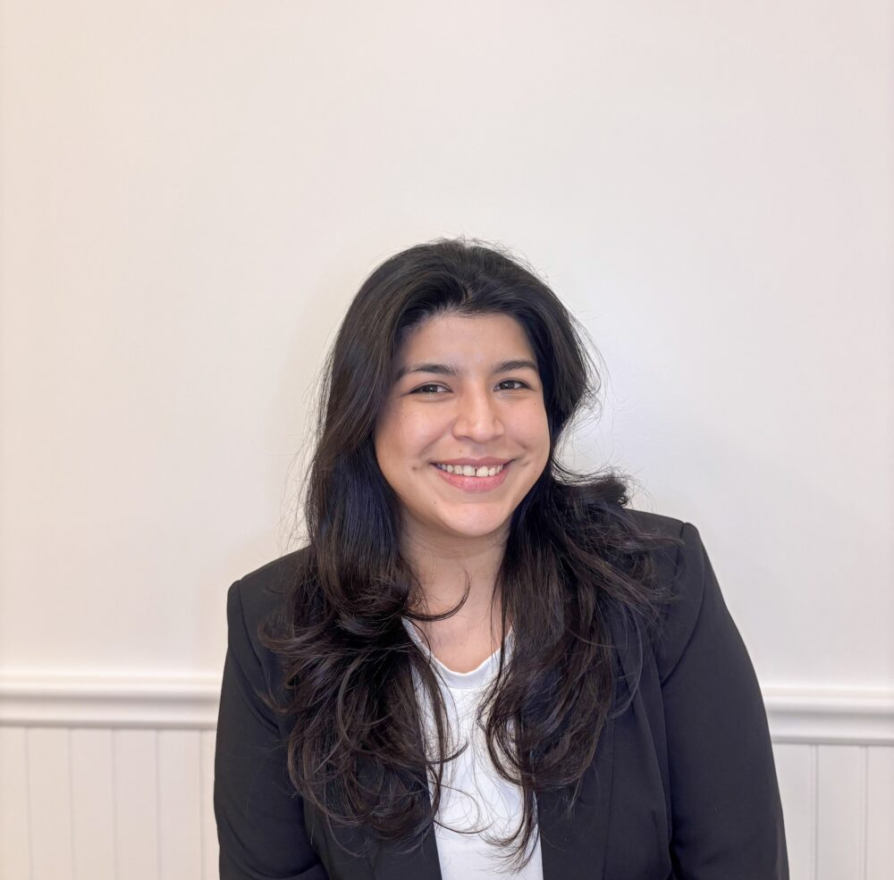 Smiling woman in a black blazer and white shirt against a light wall, cropped at chest level (professional portrait)