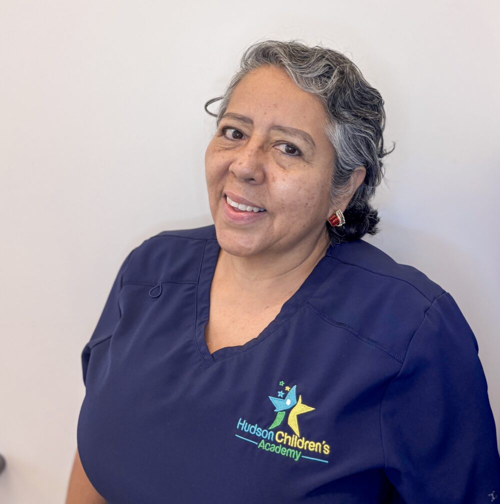 Smiling woman in navy scrubs with a small, colorful logo on the chest against a light background.