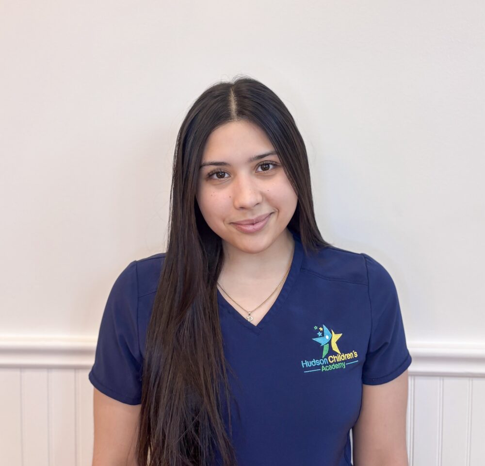 Smiling young girl in a navy polo with a colorful star logo reading Hudson Children's Academy, cropped to show shoulders and face against a light wall.