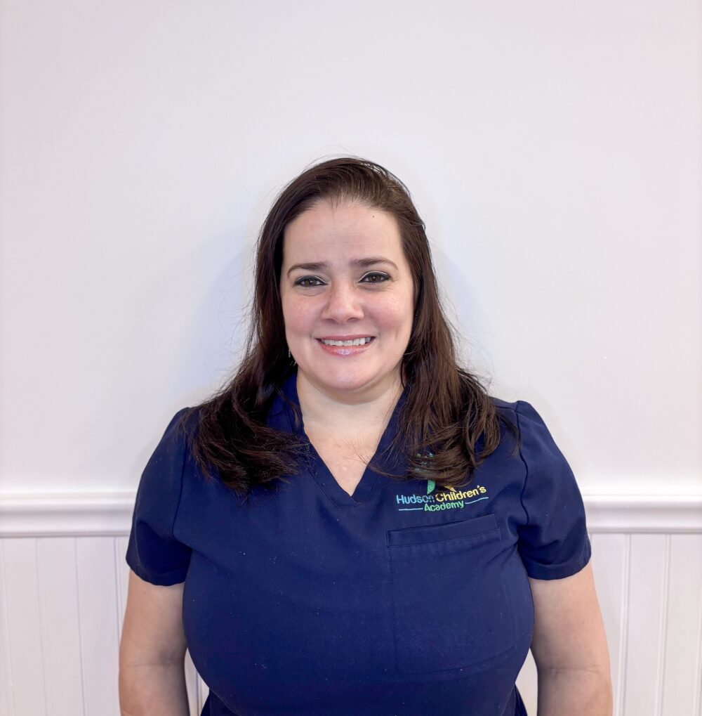Smiling woman in navy scrubs with a Hudson Children's Academy logo on the left, standing against a light wall.
