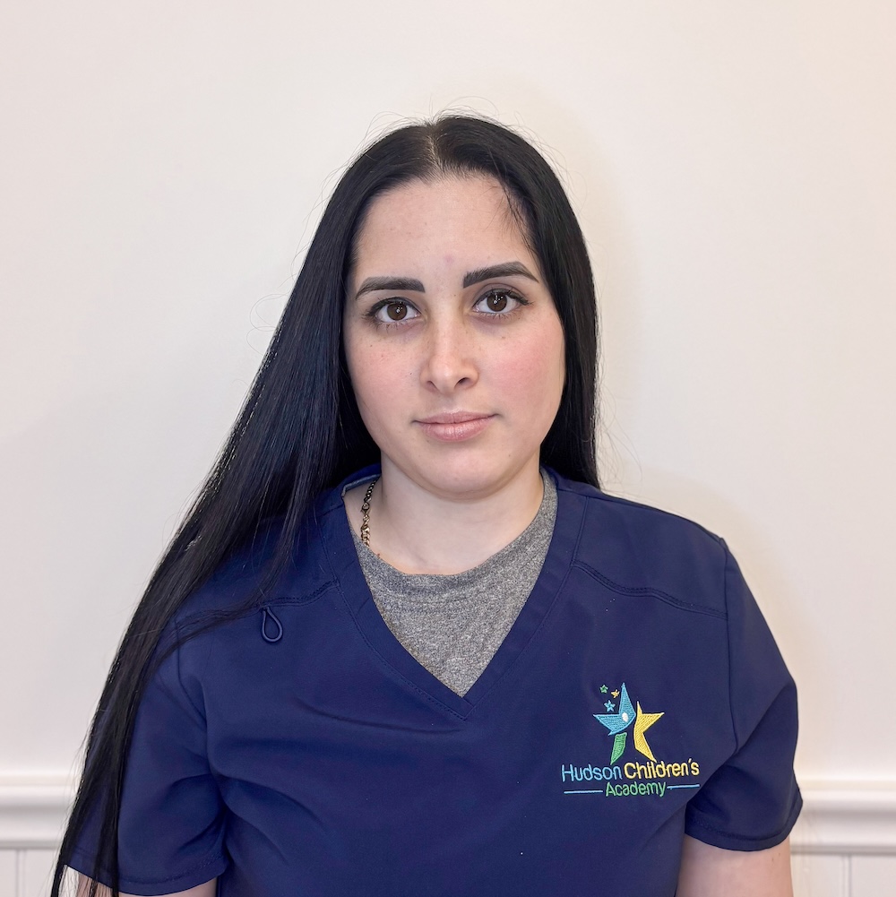 Portrait of a woman with long black hair wearing navy scrubs, facing the camera; Hudson Children’s Academy logo on her chest.