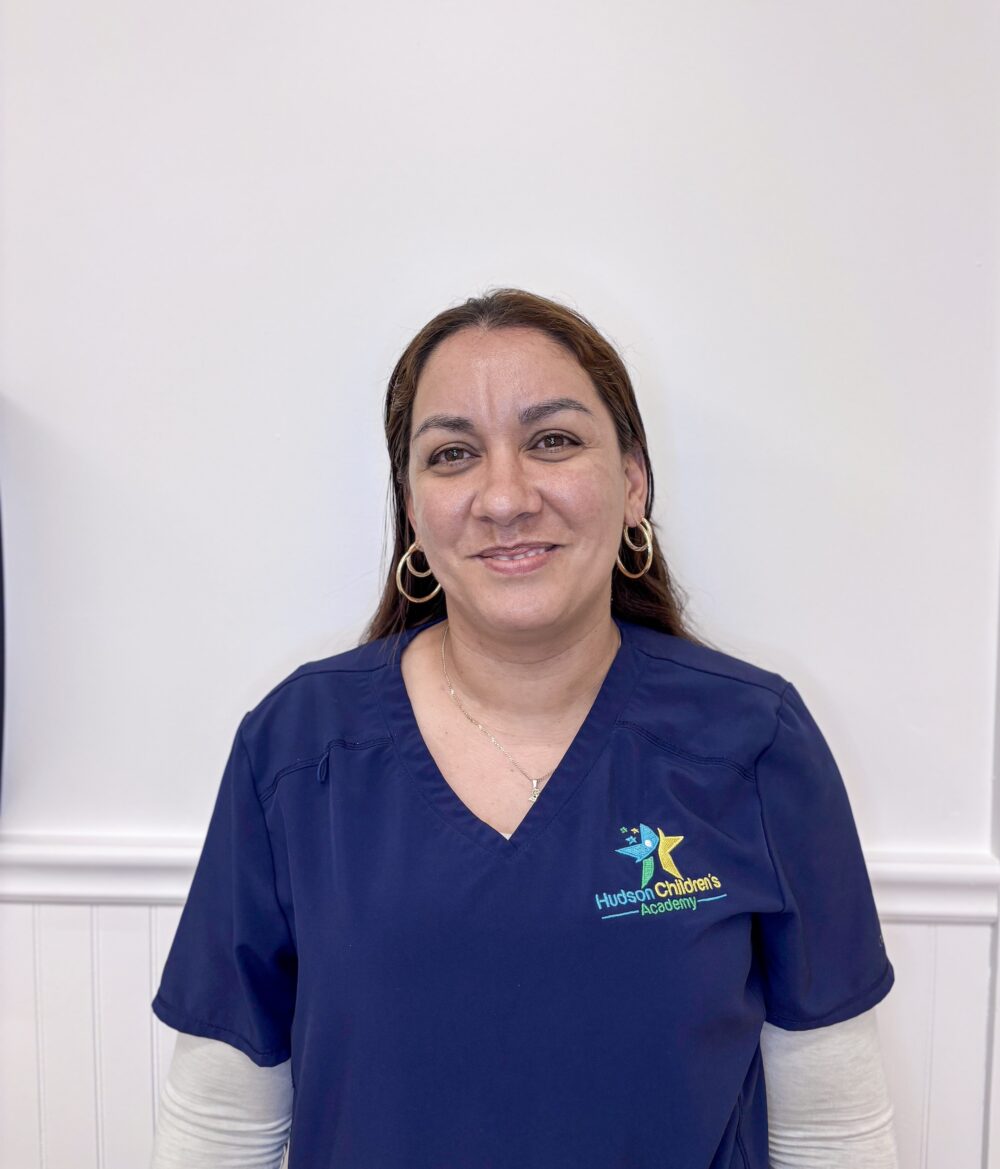 Smiling woman in navy scrubs with a logo reading 'Hudson Children's Academy' on the left chest, standing against a white wall.