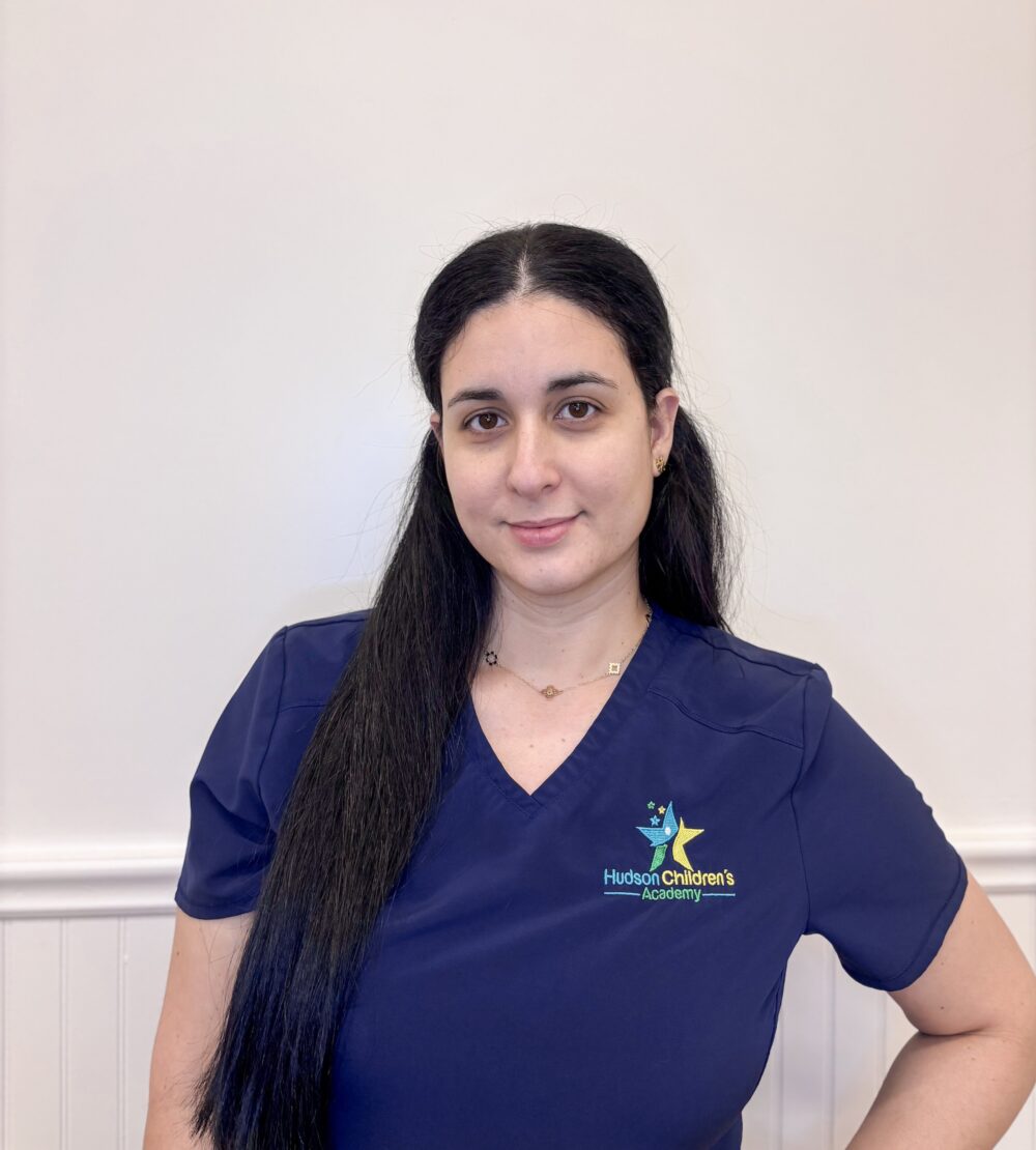 Woman in navy scrubs with Hudson Children's Academy logo on the chest, standing against a pale wall, smiling.