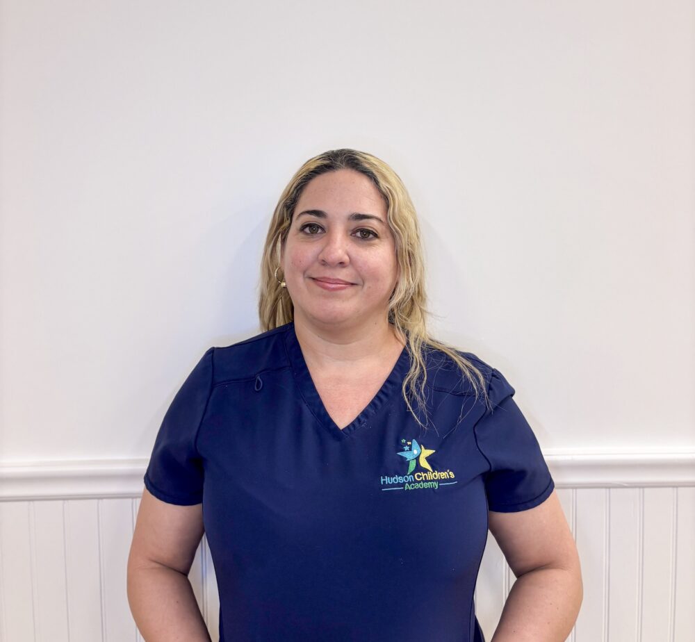 Woman in navy scrubs smiling, standing against a pale wall; Hudson Children's Academy logo on her chest.