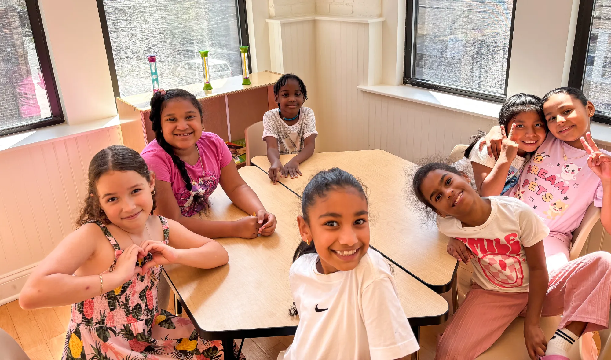 Group of smiling children gathered around a classroom table, some making peace signs, in a bright room with large windows.